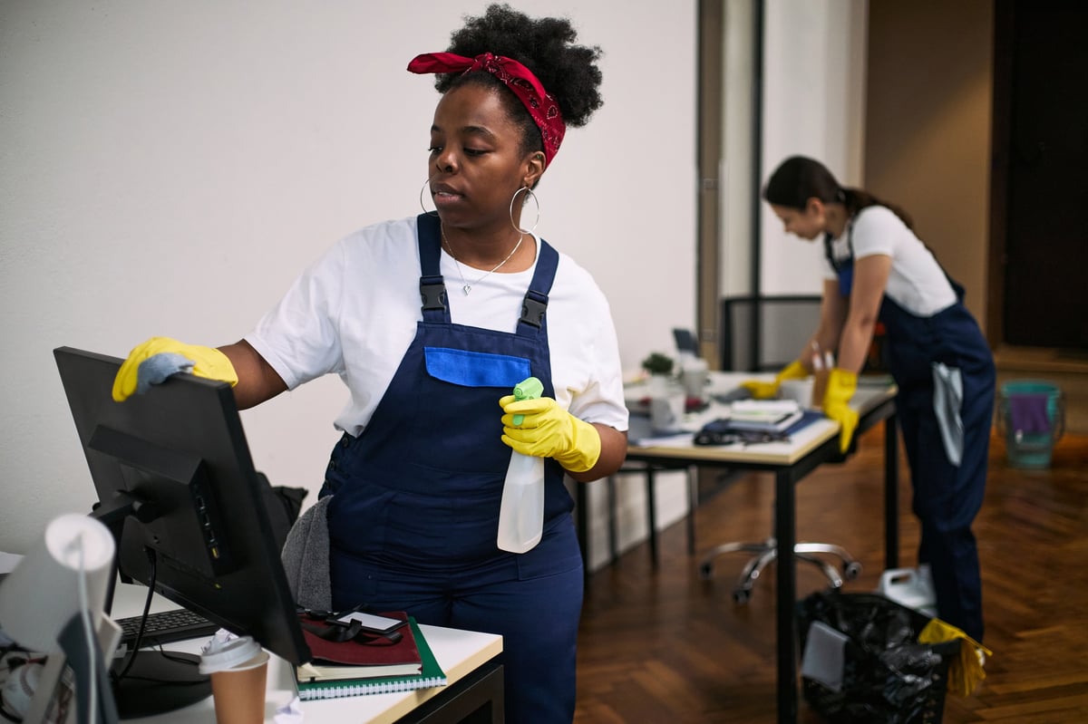Two cleaning professionals wiping down a computer monitor and organizing a desk in a modern office workspace