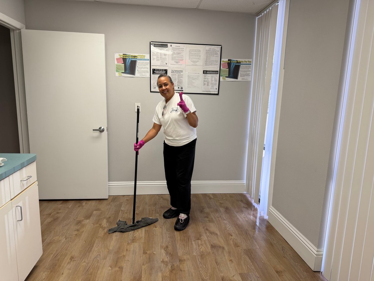 Team member in pink gloves holding a broom and dustpan on a wood floor with white walls behind her
